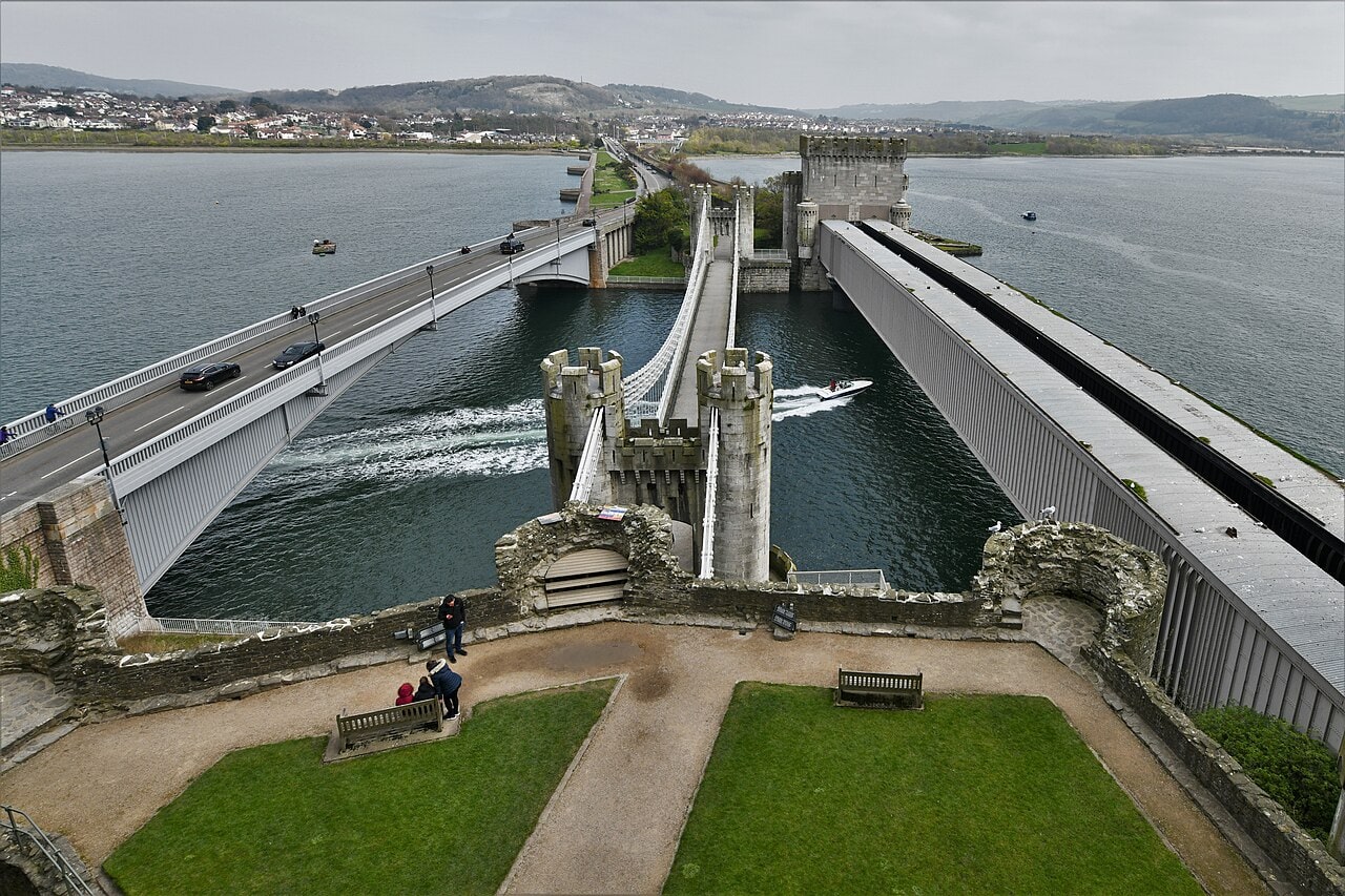 Looking down from a castle tower on a small walled garden of lawns and paths, with three bridges beyond crossing the river side by side: a road bridge, a suspension bridge between castellated towers and a boxed railway tube.