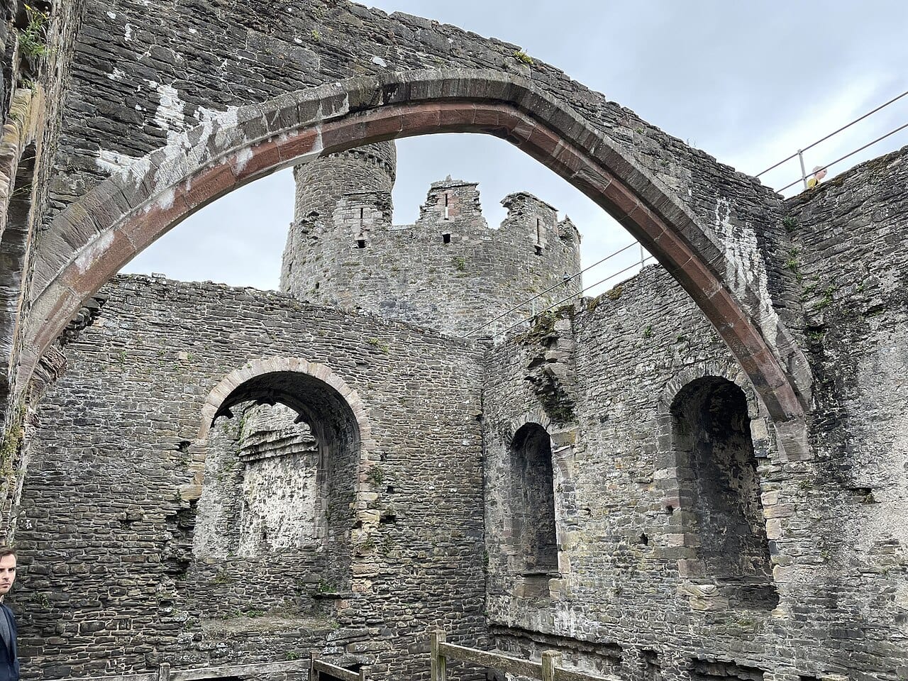 A single pointed stone arch spanning the roofless great-hall range of Conwy Castle, with round-headed window openings in the far wall and a battlemented tower behind.