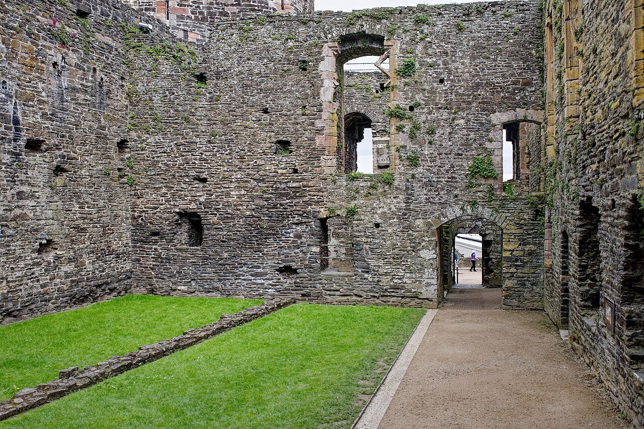 A grassy courtyard enclosed by high ruined stone walls with windows and doorways at several levels and a gravel path along one side.