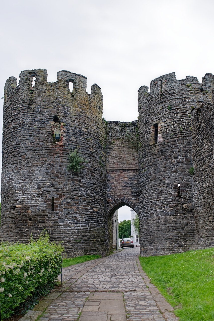 A pointed gateway arch between two battlemented stone towers in a town wall, a cobbled path leading through it.