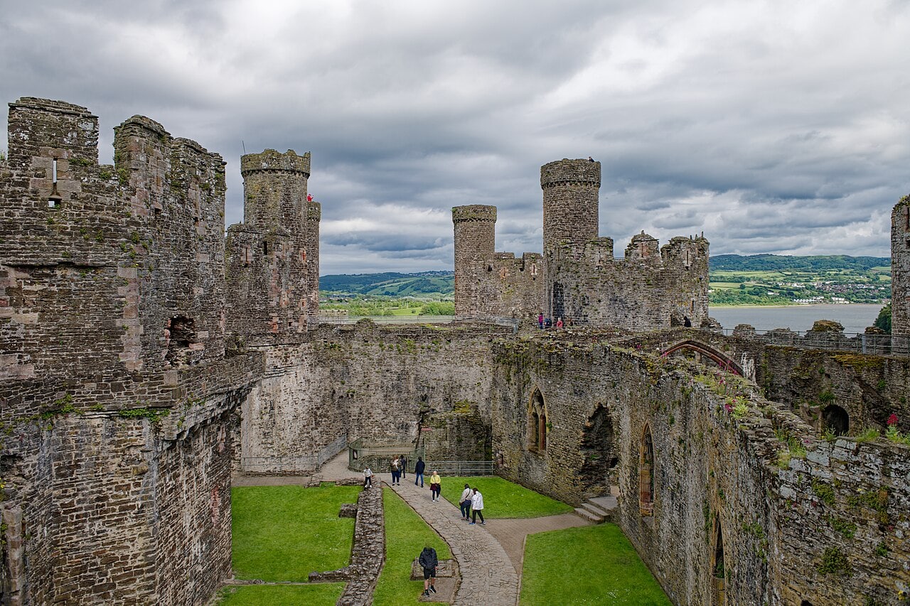 A long grassy courtyard inside Conwy Castle seen from a tower top, ruined walls with pointed windows along one side and battlemented towers with slender turrets at the far end, water beyond.