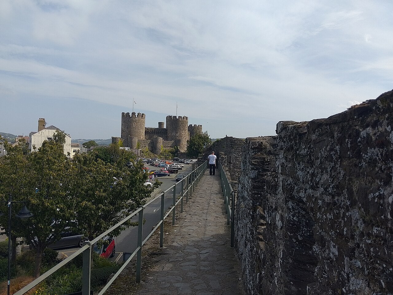 A paved walkway with a railing running along the top of a stone town wall, the towers of Conwy Castle in the distance.
