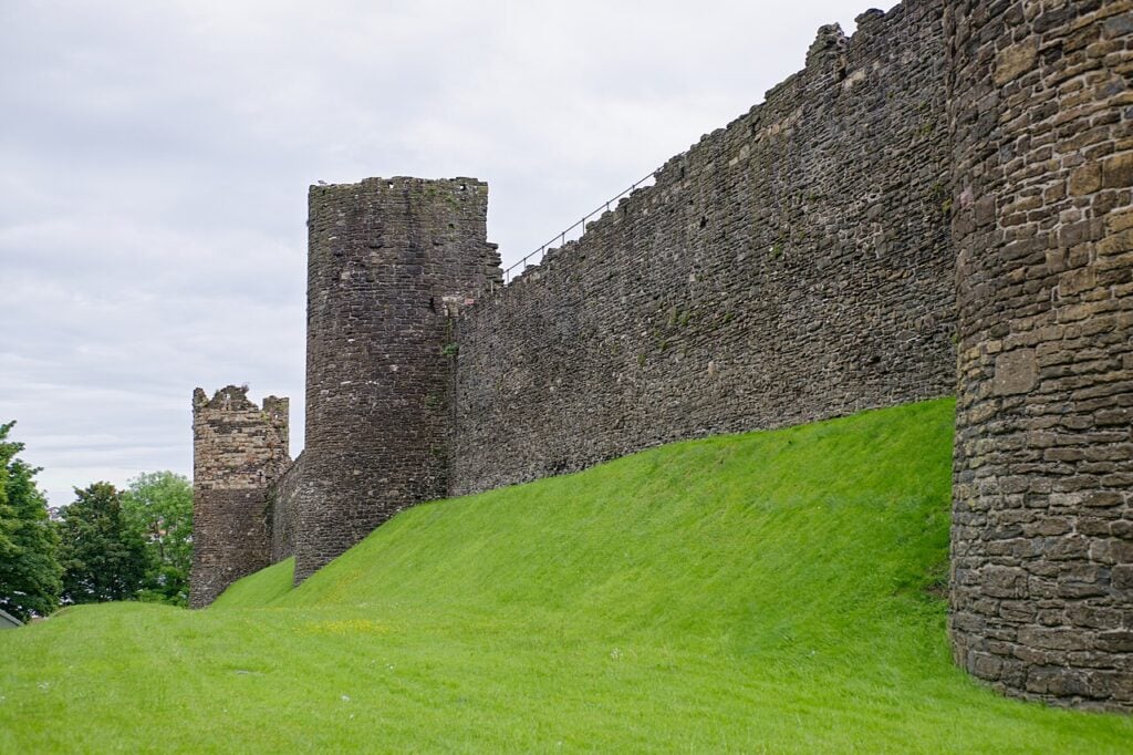 A long high stone town wall with towers rising from it above a steep green bank, the nearest tower cut off at the right edge