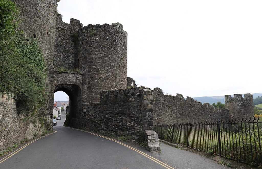 A gateway arch through a stone town wall with a tower above it, a road running through and the wall continuing downhill