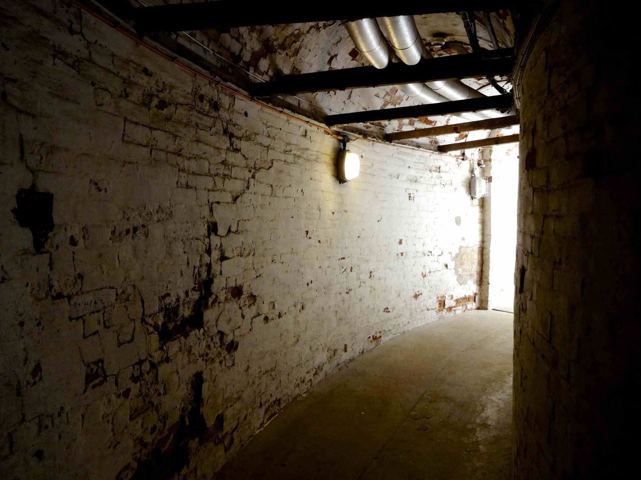 A whitewashed underground chamber with pipe and conduit runs along its ceiling and a doorway of light to the right. A whitewashed underground chamber with pipe and conduit runs along its ceiling and a doorway of light to the right.