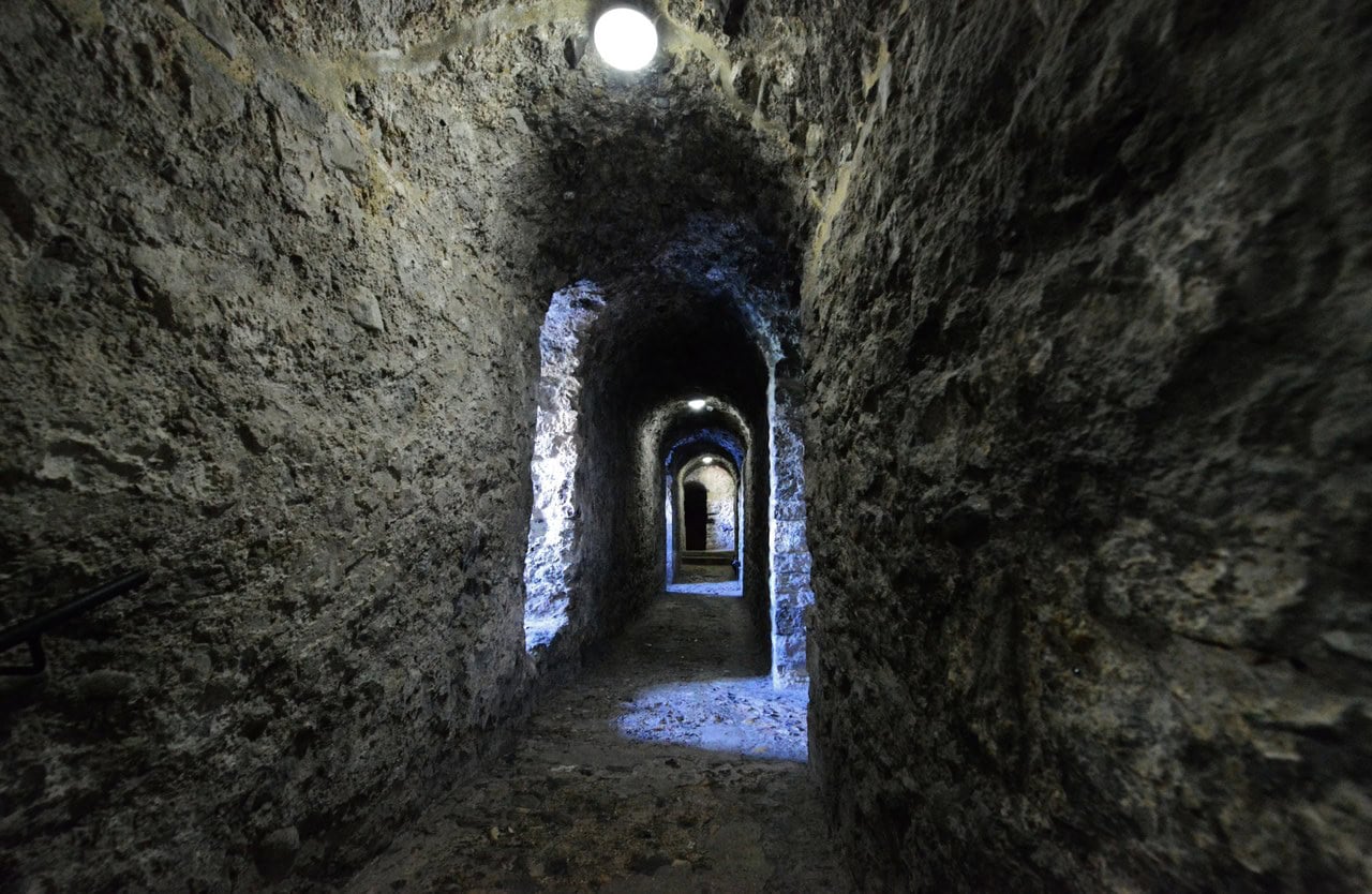 A lit stone passage running inside a thick wall, its barrel vault receding through a series of arches. A lit stone passage running inside a thick wall, its barrel vault receding through a series of arches.