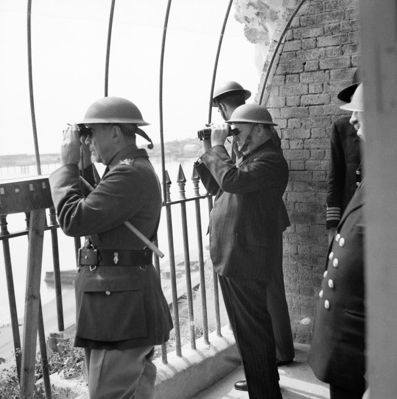Winston Churchill and uniformed officers in steel helmets looking out through binoculars from a railed opening above the water. Winston Churchill and uniformed officers in steel helmets looking out through binoculars from a railed opening above the water.