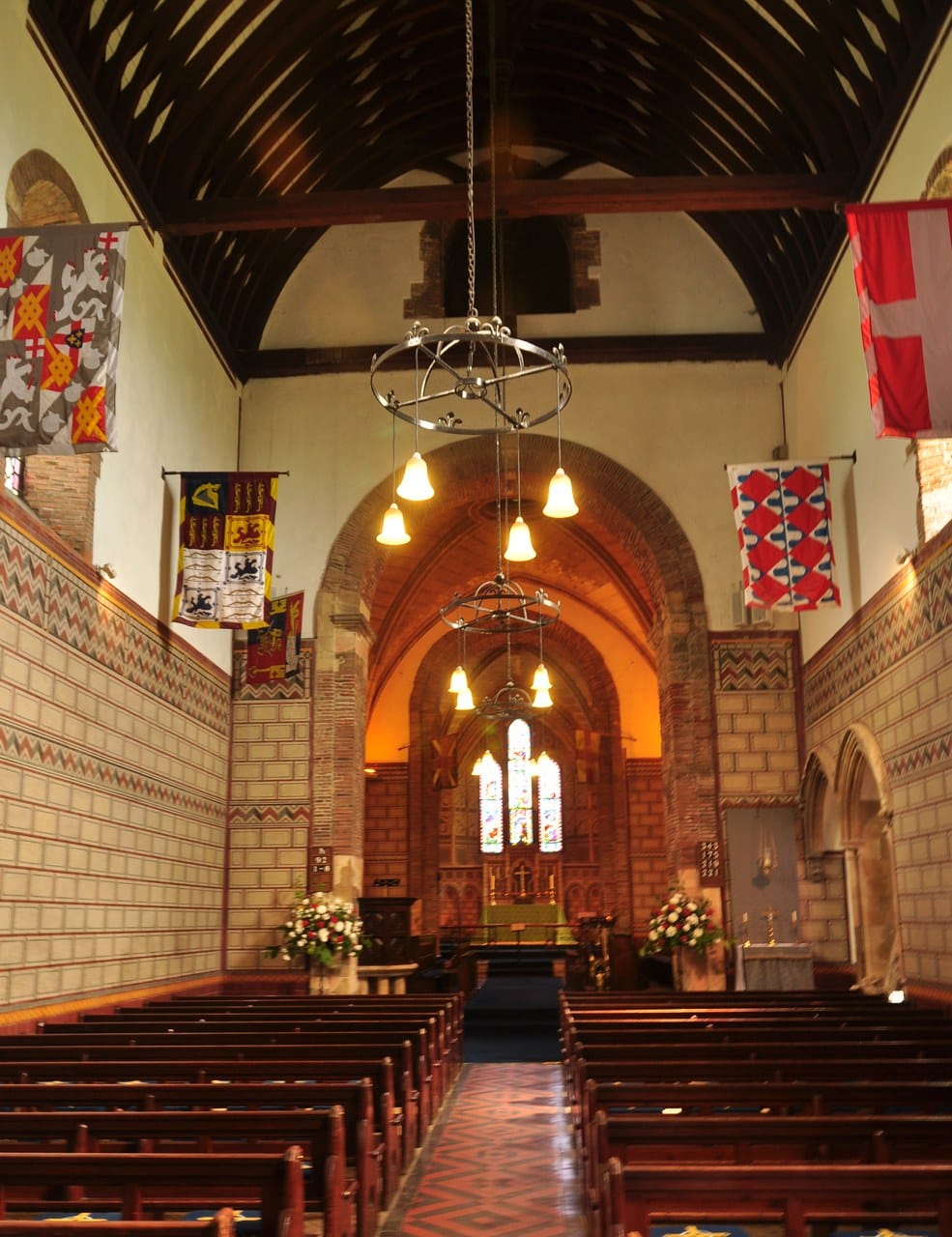 The nave of a church looking east, with hanging banners, banded painted walls and a brick arch leading to a lit chancel. The nave of a church looking east, with hanging banners, banded painted walls and a brick arch leading to a lit chancel.