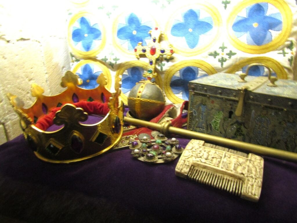 Dover Castle 12 A gilt crown, orb, sceptre, jewelled brooch, carved comb and enamelled casket displayed on purple velvet before a painted wall.
