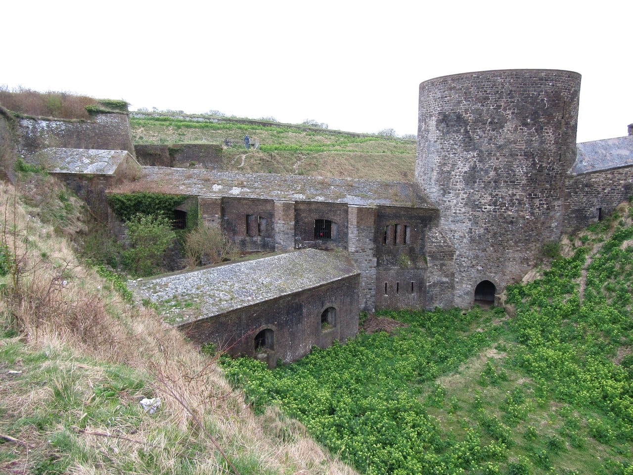 A round stone tower standing in the bottom of a grassed ditch, a stone-roofed casemate gallery running back into the bank beside it. A round stone tower standing in the bottom of a grassed ditch, a stone-roofed casemate gallery running back into the bank beside it.