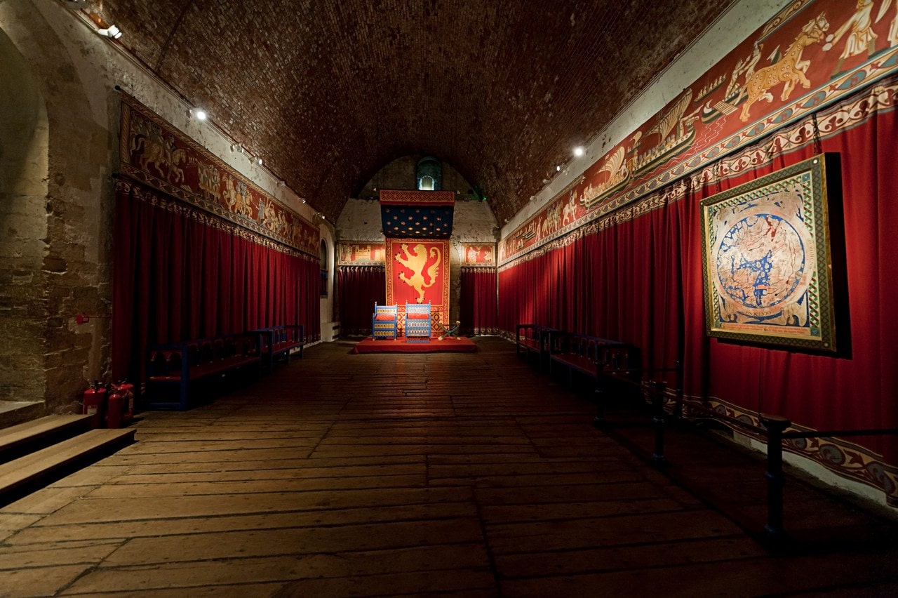 A hall hung with red drapes and painted decoration, a throne at the far end beneath a banner bearing a lion. A hall hung with red drapes and painted decoration, a throne at the far end beneath a banner bearing a lion.