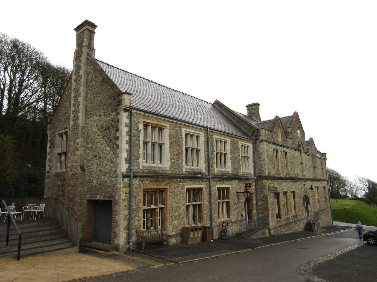 A two-storey flint and stone range with mullioned windows and a slate roof, cafe tables set out on a railed terrace at the left. A two-storey flint and stone range with mullioned windows and a slate roof, cafe tables set out on a railed terrace at the left.