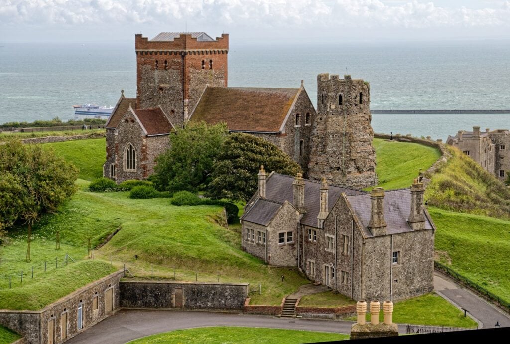 Dover Castle 7 The ruined Roman pharos beside the flint church of St Mary in Castro on high ground, with a stone range below and the sea behind.