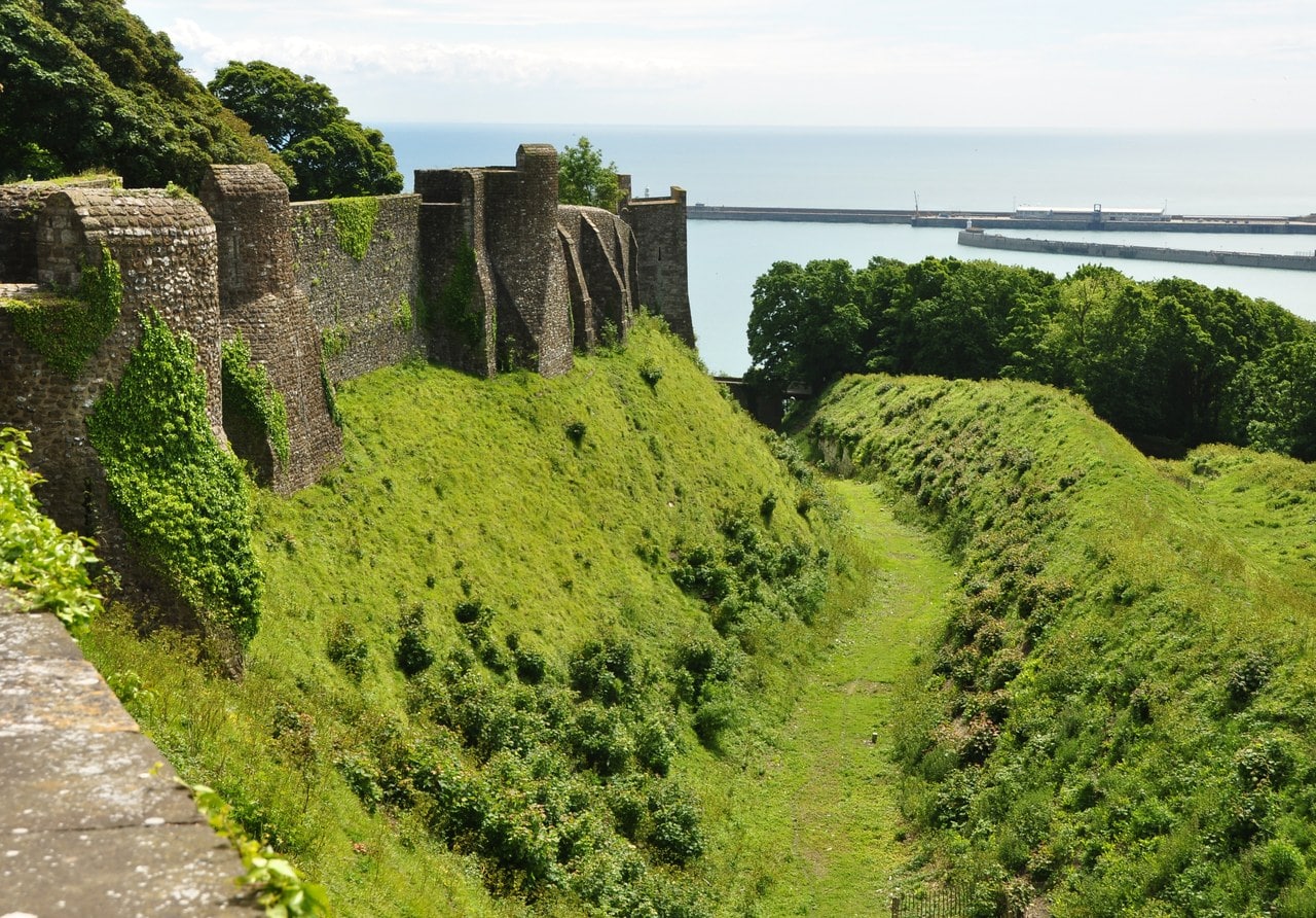 A deep grassed ditch curving beneath the castle's curtain wall, with the sea on the horizon. A deep grassed ditch curving beneath the castle's curtain wall, with the sea on the horizon.