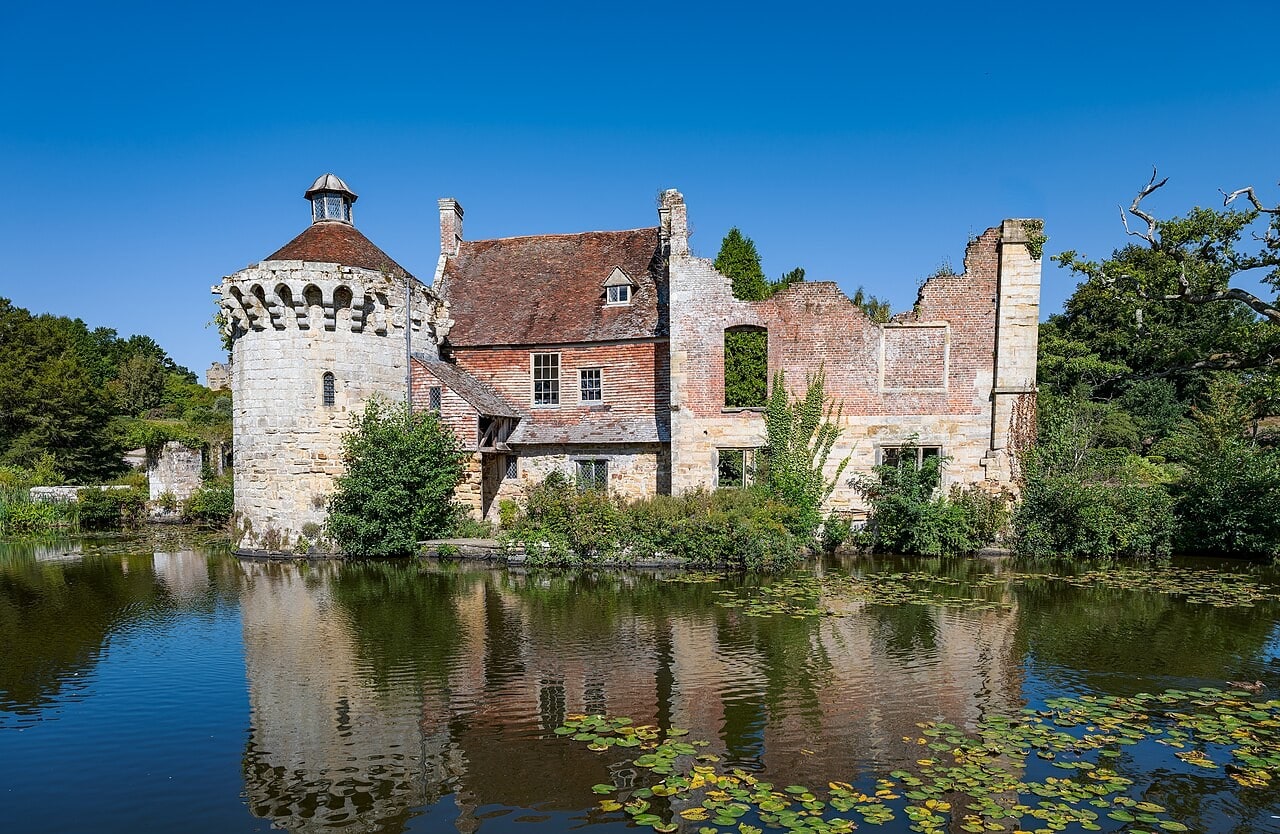 A round stone tower with a small domed lantern beside a red-brick house with a tiled roof and a roofless ruined wing to its right, all reflected in a still moat under a clear blue sky. A round stone tower with a small domed lantern beside a red-brick house with a tiled roof and a roofless ruined wing to its right, all reflected in a still moat under a clear blue sky.