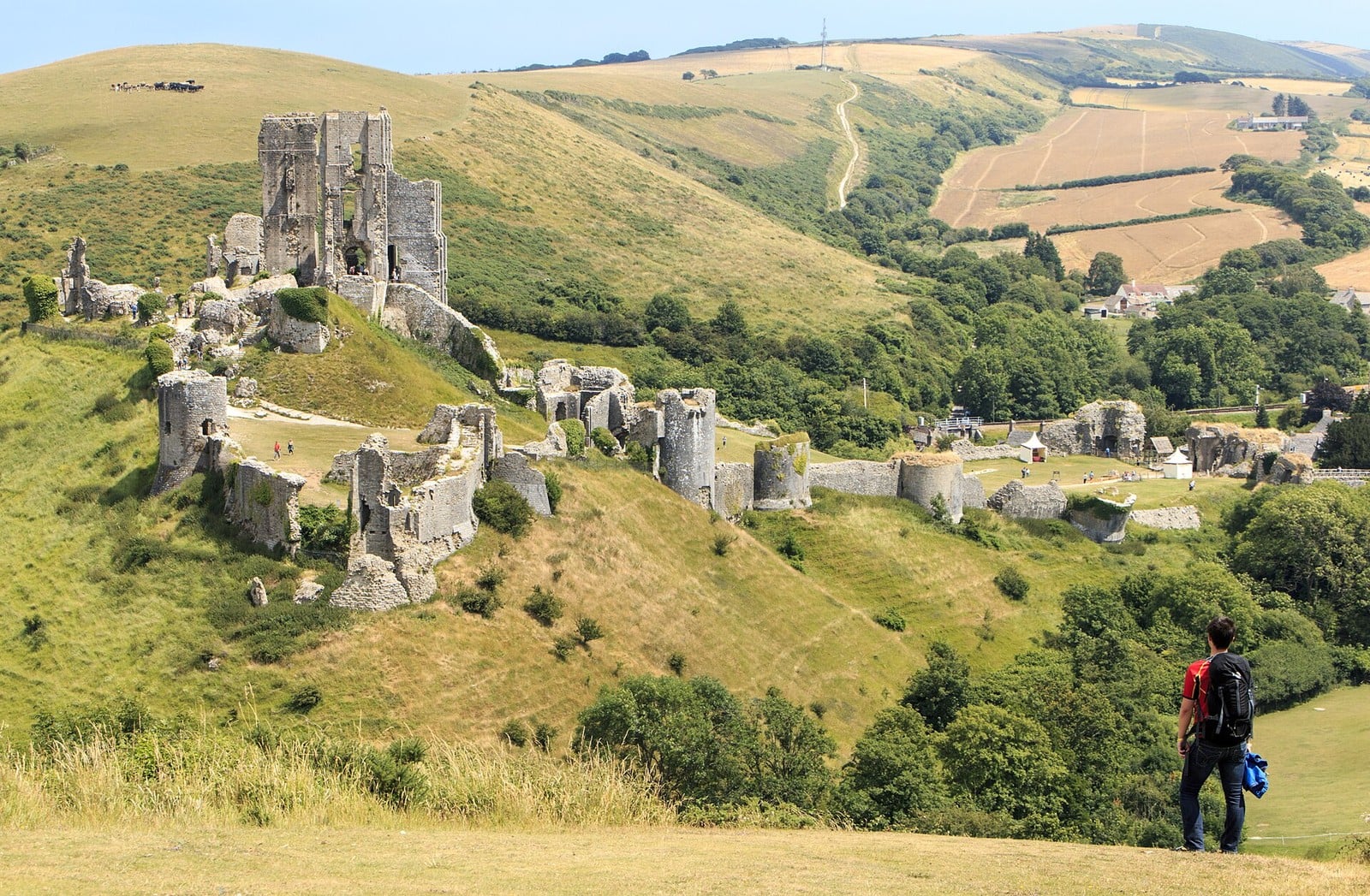 Why Are English Castles in Ruins? 1 The ruins of Corfe Castle spread across a steep green hill: a tall broken keep at the top, ruined towers and lengths of wall scattered down the slope, fields and hills behind, and a hiker with a backpack standing in the right foreground.