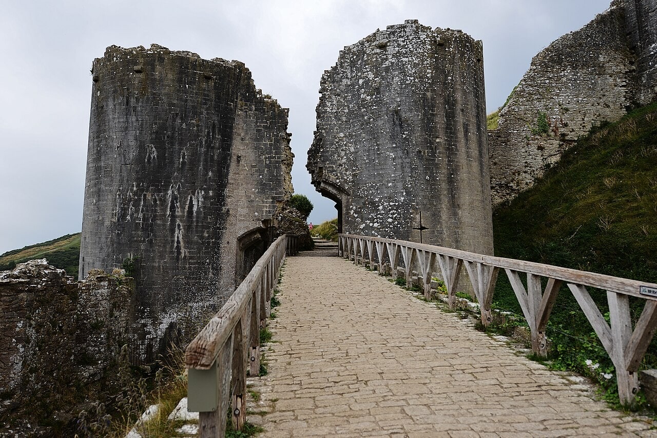 A paved bridge with timber railings leading to a gateway between two tall round stone towers that stand apart, the gap between them open to the sky where the wall over the gate has broken away. A paved bridge with timber railings leading to a gateway between two tall round stone towers that stand apart, the gap between them open to the sky where the wall over the gate has broken away.