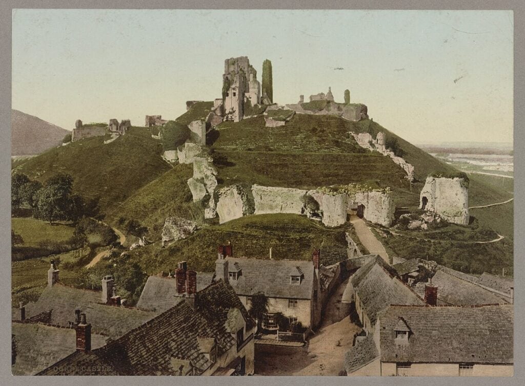 Why Are English Castles in Ruins? 2 A colored photochrom print of a ruined castle spread over a green hill above the gray stone roofs of a village, with a wide plain beyond.