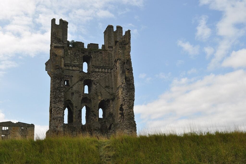 Why Are English Castles in Ruins? 9 A tall ruined stone tower with a battlemented top, its near face opened up to show window openings on several floors, rising from a grassy bank under a blue sky with light clouds.