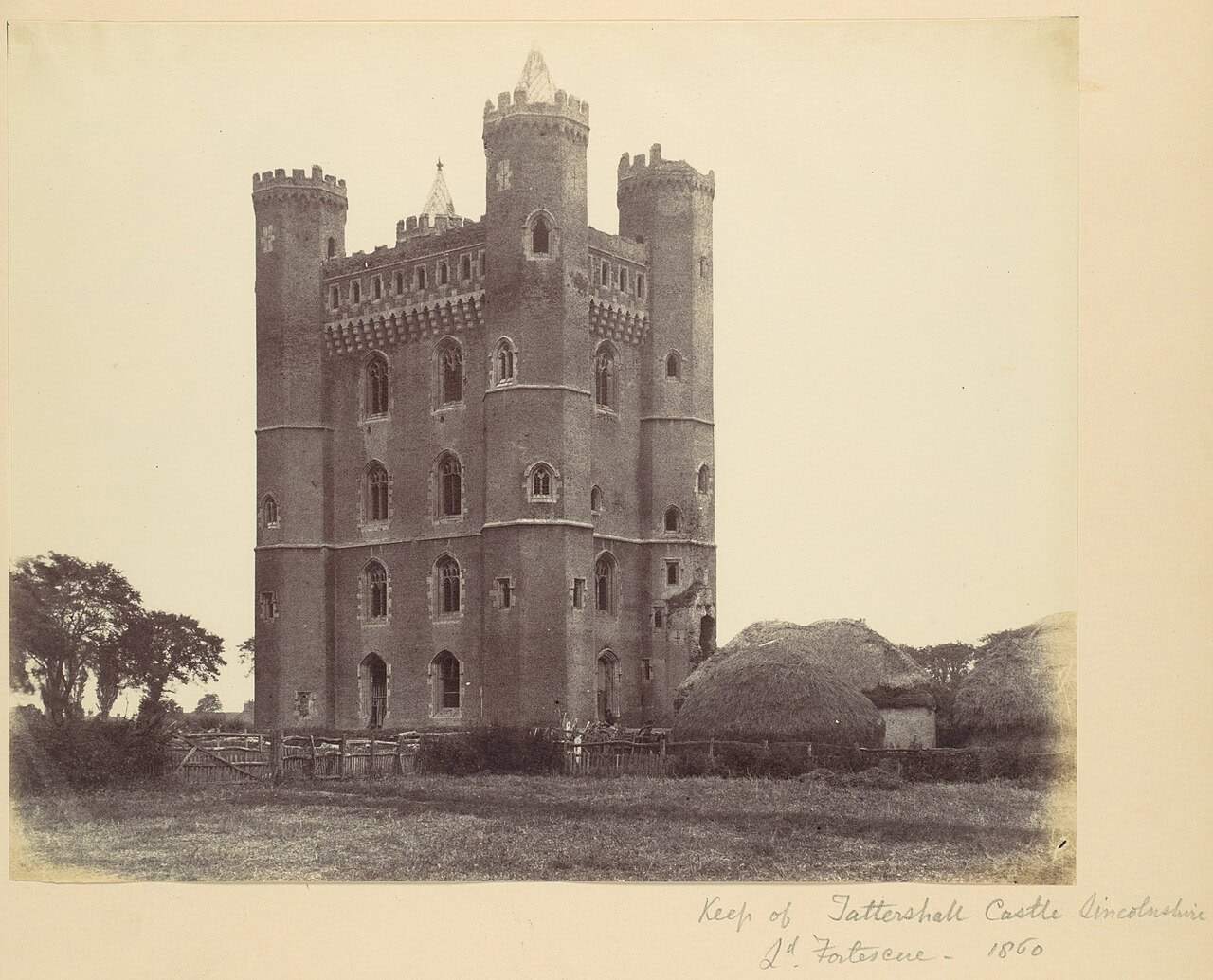 A sepia photograph of a tall brick tower with four corner turrets and rows of windows, standing in open ground beside haystacks and trees, mounted on card with a handwritten inscription below. A sepia photograph of a tall brick tower with four corner turrets and rows of windows, standing in open ground beside haystacks and trees, mounted on card with a handwritten inscription below.