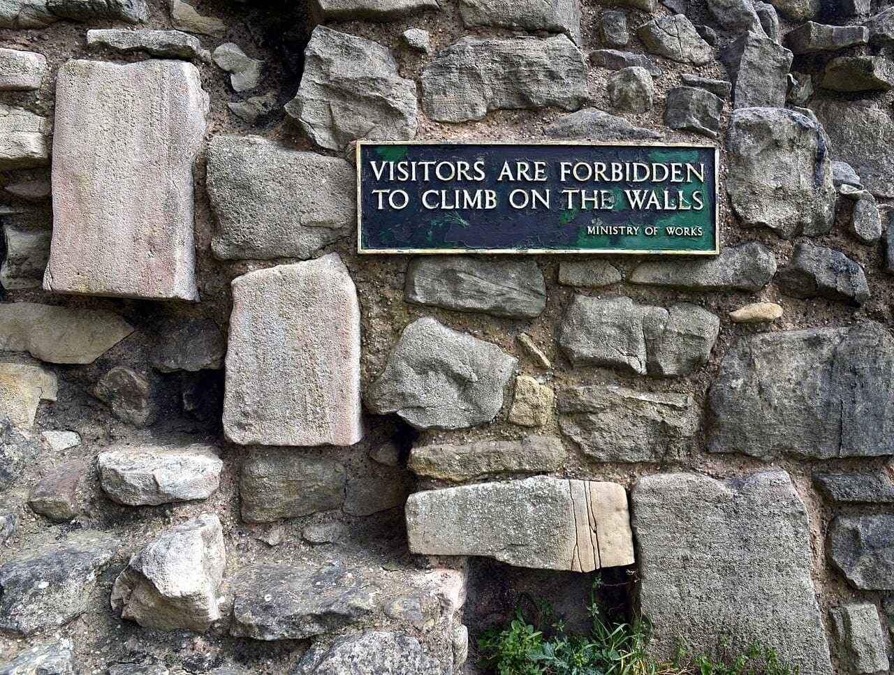 A small green metal sign reading VISITORS ARE FORBIDDEN TO CLIMB ON THE WALLS, with MINISTRY OF WORKS in small letters beneath, fixed to rough stone masonry. A small green metal sign reading VISITORS ARE FORBIDDEN TO CLIMB ON THE WALLS, with MINISTRY OF WORKS in small letters beneath, fixed to rough stone masonry.
