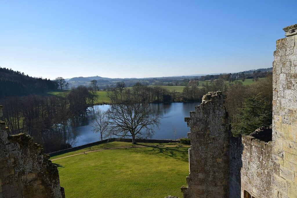 Why Are English Castles in Ruins? 8 Looking out between broken stone walls over a lawn, woodland and a lake in a wide wooded valley under a clear blue sky.