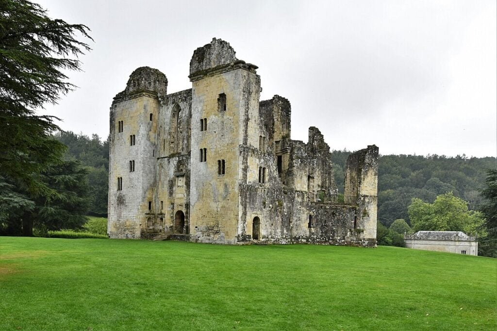 Why Are English Castles in Ruins? 7 A tall pale stone castle with a many-sided plan and rows of empty windows, one part standing to full height and another broken down, on a lawn with dark trees at the left.