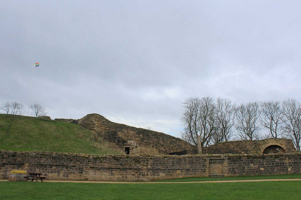 A long low stone wall across a lawn under a gray sky, with a grassed mound topped by a flagpole, a curved mass of ruined masonry, bare trees and a low arched ruin beyond. A long low stone wall across a lawn under a gray sky, with a grassed mound topped by a flagpole, a curved mass of ruined masonry, bare trees and a low arched ruin beyond.