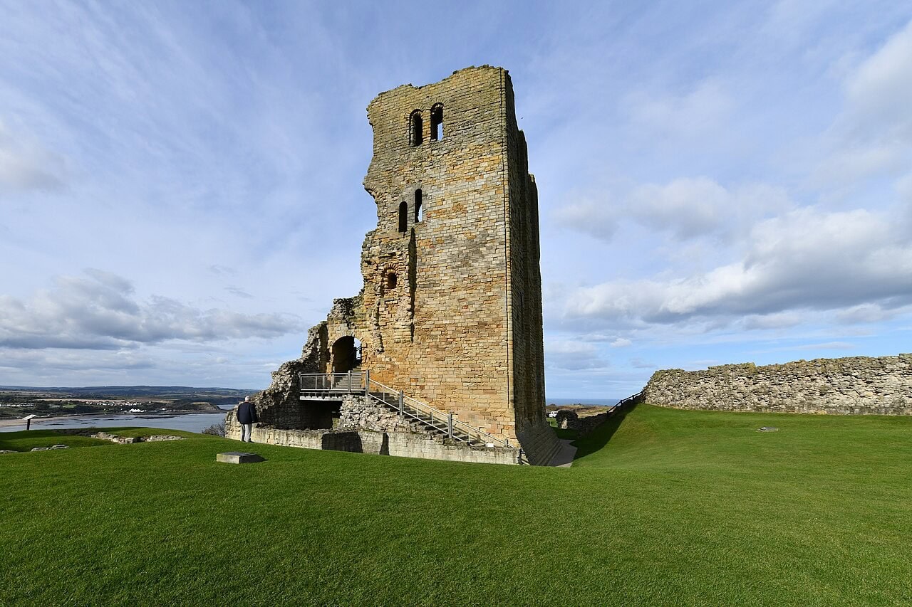 A tall rectangular stone tower standing to its full height on one side only, its broken edge stepping down to a ruined base with a timber stair and platform, on a wide lawn with a low wall and water beyond. A tall rectangular stone tower standing to its full height on one side only, its broken edge stepping down to a ruined base with a timber stair and platform, on a wide lawn with a low wall and water beyond.