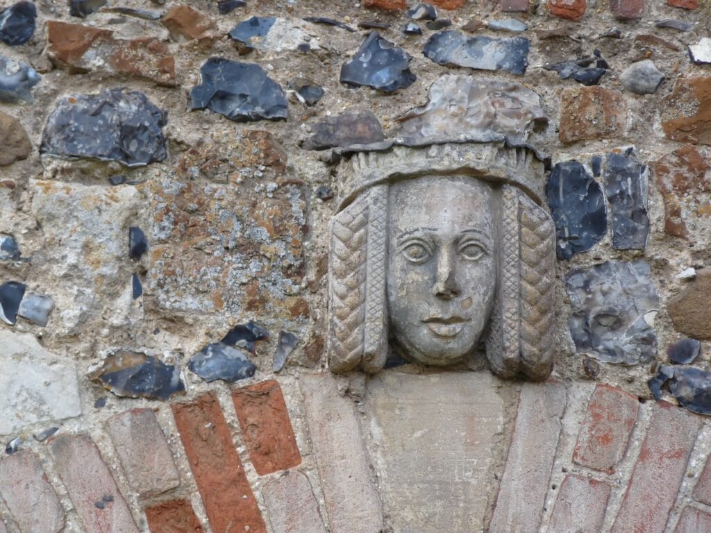 Framlingham Castle 10 A carved stone head set into a castle wall. The face is that of a figure wearing a crown or band across the brow, with long hair falling in plaited strands on either side. The surrounding wall is built of knapped flint in blue and gray with bands of red brick below.