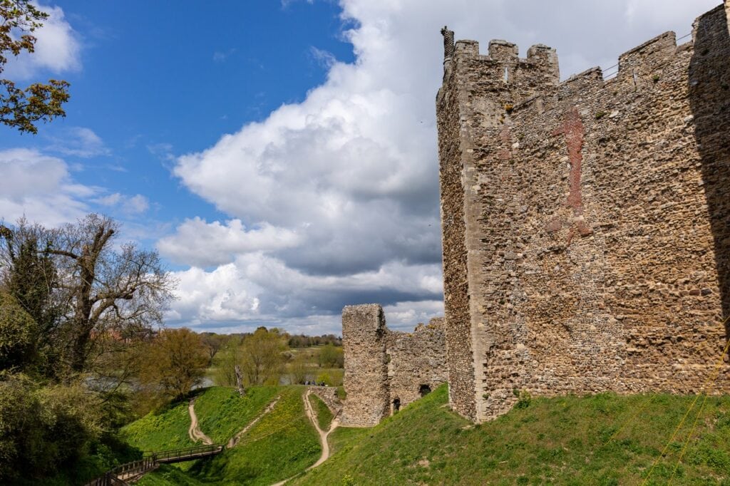 Framlingham Castle 4 A tall square castle tower rising from a steep grassed bank, seen from outside on a bright day. The tower is built of flint rubble with dressed stone at its corners and a crenellated top, and a lower stretch of curtain wall runs away from it to the left. Footpaths cross the green slope below. Trees stand at the left with a glimpse of water and open ground beyond. Blue sky with large white clouds.
