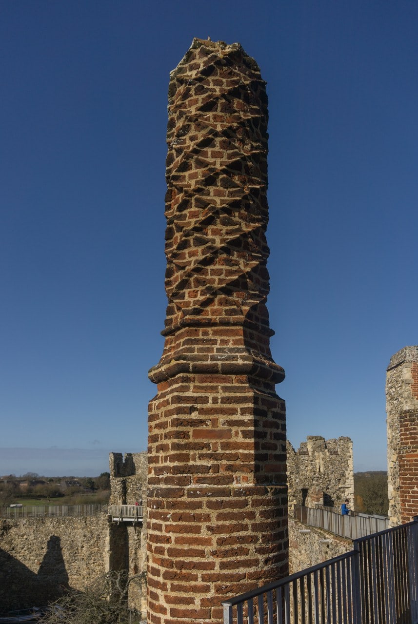 A tall cylindrical chimney rising from the castle wall-walk against a clear blue sky. The lower part is a smooth round drum of coursed brickwork. A molded collar marks a clear horizontal junction partway up, and above it the shaft is carried up in brickwork laid in a twisting spiral. Behind and below, the crenellated curtain wall, a metal handrail and open countryside can be seen. A tall cylindrical chimney rising from the castle wall-walk against a clear blue sky. The lower part is a smooth round drum of coursed brickwork. A molded collar marks a clear horizontal junction partway up, and above it the shaft is carried up in brickwork laid in a twisting spiral. Behind and below, the crenellated curtain wall, a metal handrail and open countryside can be seen.