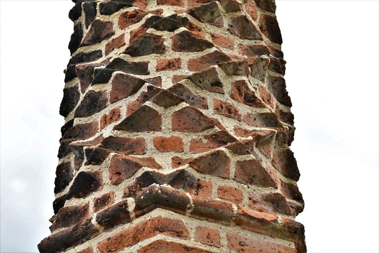 A close view of a chimney shaft built of red brick, filling the frame against a pale overcast sky. The bricks are laid so that projecting darker headers form a bold repeating zig-zag or chevron pattern running around the shaft, with pale mortar joints between them. A close view of a chimney shaft built of red brick, filling the frame against a pale overcast sky. The bricks are laid so that projecting darker headers form a bold repeating zig-zag or chevron pattern running around the shaft, with pale mortar joints between them.