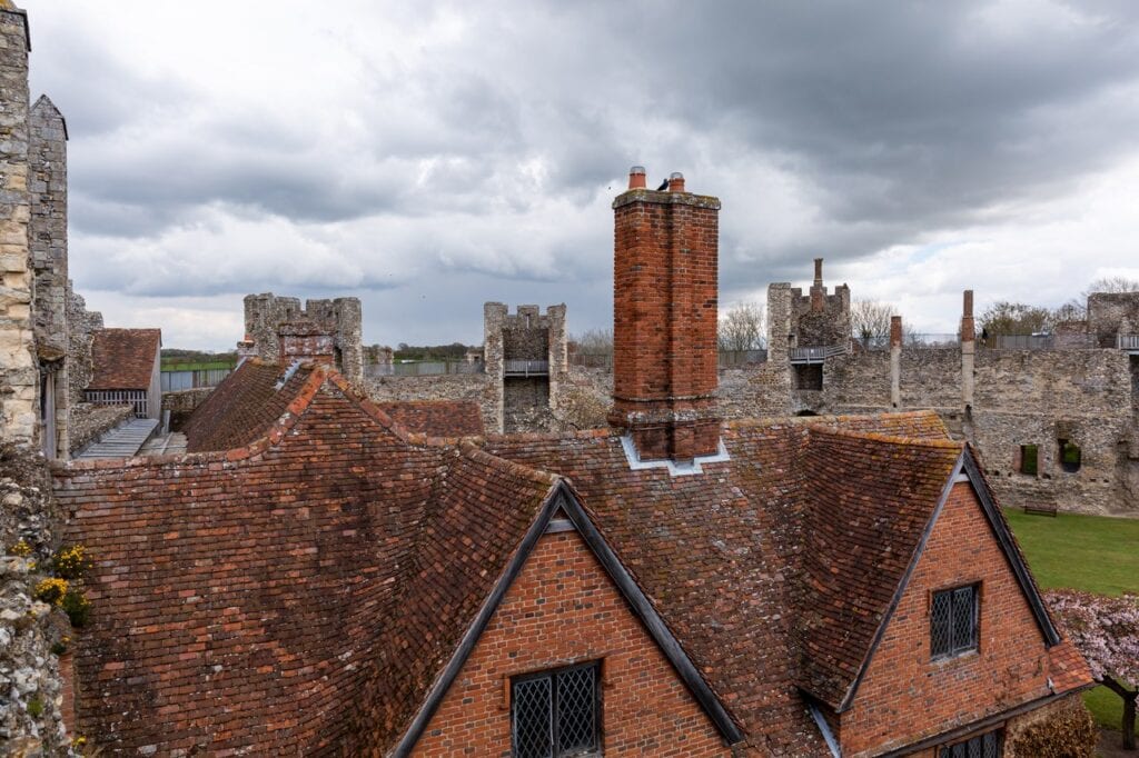 Framlingham Castle 9 A view across the red tiled roofs of the range inside the castle, taken from above. A large red brick chimney stack rises in the center of the frame, with dormer windows and a gabled roof below it. The crenellated flint curtain wall and further chimney shafts stand behind under a heavy gray sky.