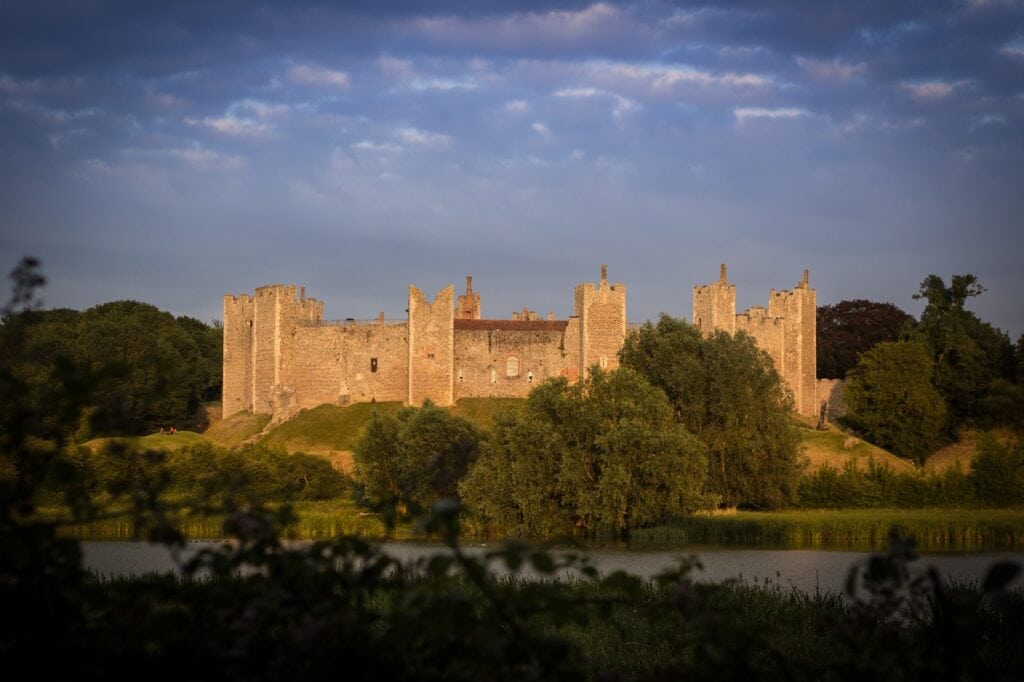 Framlingham Castle 2 Framlingham Castle at sunset. The curtain wall and its square towers are lit gold by low sun against a deep blue sky streaked with cloud. Trees in full summer leaf stand along the foot of the grassy bank below the wall. Across the lower part of the frame lies a still sheet of water with a reed fringe, seen past dark out-of-focus foliage in the immediate foreground.