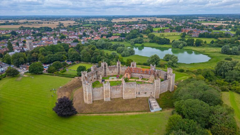 Home 7 An aerial view of Framlingham Castle. A continuous gray stone curtain wall runs in a rough oval around a level green court, with square towers standing at intervals along it and slender chimney shafts rising from several of them. Inside the wall the ground is almost entirely open grass; a single long building with a red tiled roof stands against the far side. A broad grassed bank falls away outside the wall, with mown lawn beyond it. The town of Framlingham fills the left of the frame, a church tower among its roofs. On the right, low green meadow stretches away with one irregular pool of open water lying in it, fringed by trees. Flat farmland reaches to the horizon under a cloudy sky.