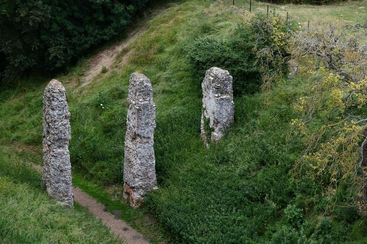 Three tall weathered stone piers standing in a line in a grassy ditch, each roughly rectangular and built of pale rubble, rising to about head height. A narrow worn footpath runs between them. Steep green banks covered in grass, brambles and yellow flowers rise on either side, with a wire fence and trees along the top. Three tall weathered stone piers standing in a line in a grassy ditch, each roughly rectangular and built of pale rubble, rising to about head height. A narrow worn footpath runs between them. Steep green banks covered in grass, brambles and yellow flowers rise on either side, with a wire fence and trees along the top.