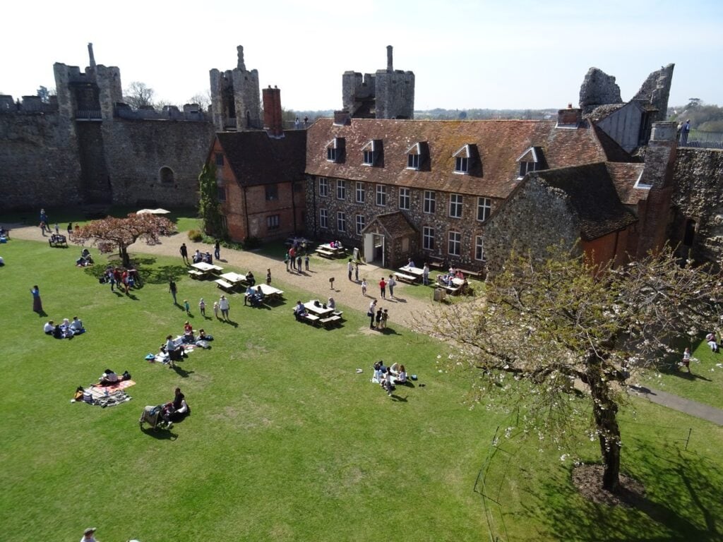 Framlingham Castle 8 The inner court of the castle seen from above on a sunny spring day. A long building with a red tiled roof and dormer windows stands along the far side, with cars parked in front of it and visitors sitting and walking on the wide green lawn. The crenellated curtain wall and its square towers enclose the court. A tree in white blossom stands at the right.
