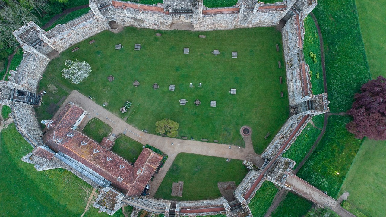A near-vertical aerial view looking straight down into Framlingham Castle. The curtain wall reads as a closed ring of pale masonry with its towers projecting outward as square blocks. The enclosure inside is almost all green lawn with scattered benches and picnic tables and a gravel path curving across it. One long building with a red tiled roof stands along the lower left side of the ring. Outside the wall the grassed ditch and bank can be seen, with a copper-leaved tree at the right. A near-vertical aerial view looking straight down into Framlingham Castle. The curtain wall reads as a closed ring of pale masonry with its towers projecting outward as square blocks. The enclosure inside is almost all green lawn with scattered benches and picnic tables and a gravel path curving across it. One long building with a red tiled roof stands along the lower left side of the ring. Outside the wall the grassed ditch and bank can be seen, with a copper-leaved tree at the right.