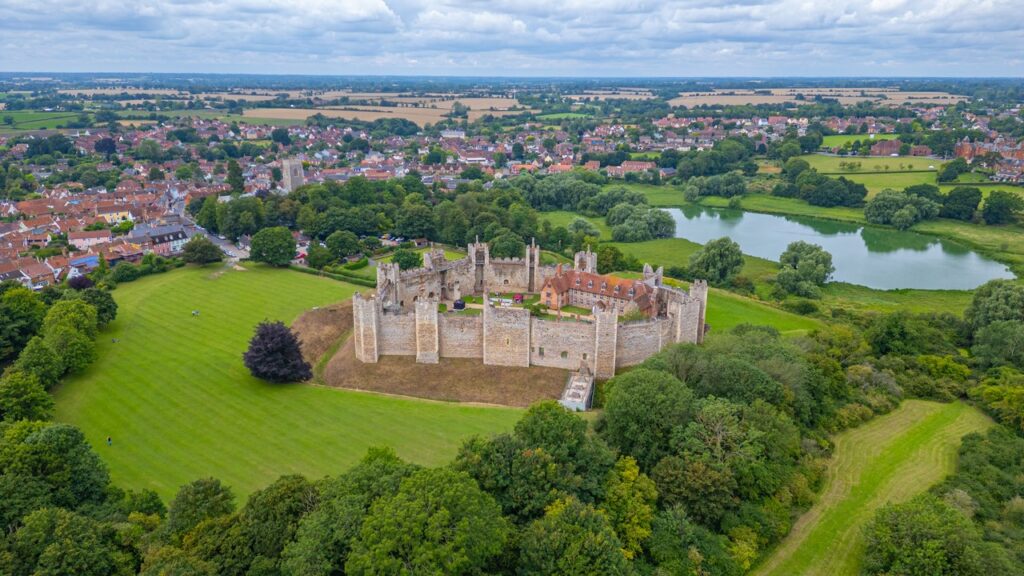 Framlingham Castle 3 An aerial view of Framlingham Castle in its setting. The oval stone curtain wall with its projecting square towers encloses a green court containing one long red-roofed building. The town of Framlingham, with a church tower, occupies the left half of the frame. To the right and below, broad green meadow is divided by tree lines and holds a single irregular pool of open water. Mown parkland, woodland and flat farmland extend to the horizon.