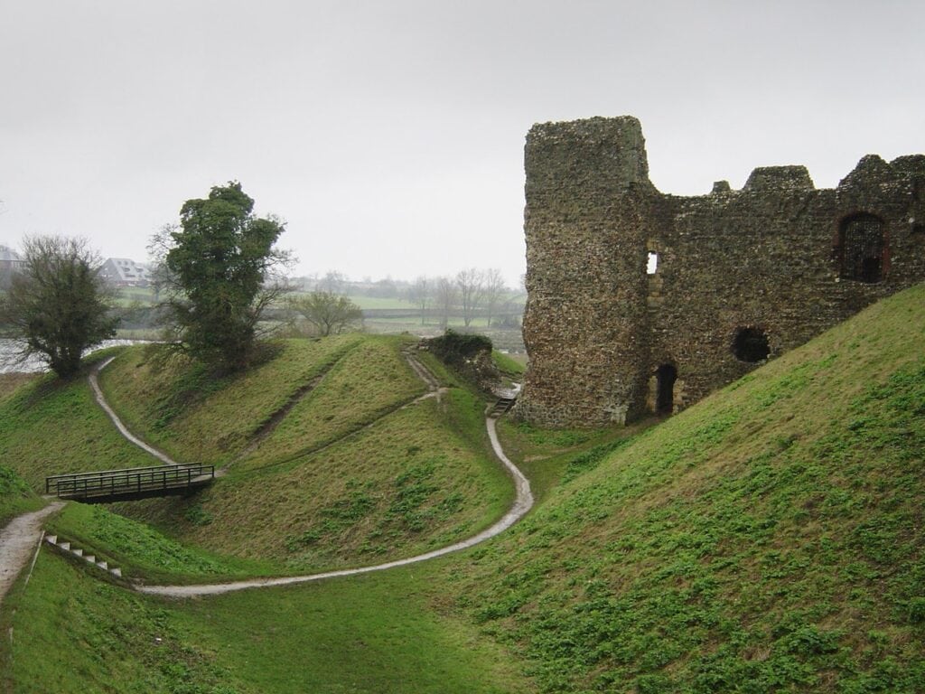 Framlingham Castle 11 The dry ditch at the foot of the castle on a gray day. Smooth green banks fall steeply to the ditch floor, with narrow footpaths worn across them and a small wooden footbridge spanning the hollow at the left. A tall fragment of ruined flint wall stands on the bank at the right, with square openings in its face. Bare and lightly leafed trees stand around, with flat fields and a pale sky beyond.