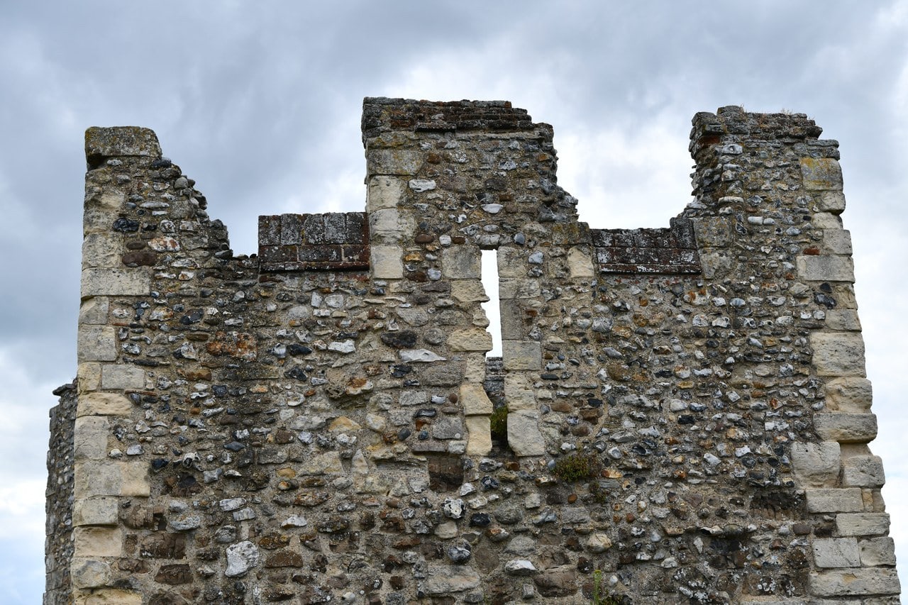 The upper part of a square castle tower seen from outside against a gray cloudy sky. Its side walls stand to full height with dressed stone quoins at the corners and flint rubble walling between, and the parapet is crenellated. The back of the tower is open, so daylight shows straight through the gap where a rear wall would be. The upper part of a square castle tower seen from outside against a gray cloudy sky. Its side walls stand to full height with dressed stone quoins at the corners and flint rubble walling between, and the parapet is crenellated. The back of the tower is open, so daylight shows straight through the gap where a rear wall would be.