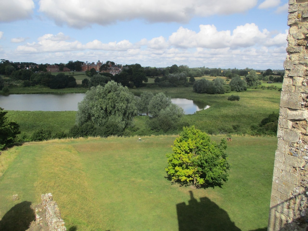 A view outward from the castle wall, taken over the parapet with masonry at the right edge. Below lies a wide area of green meadow crossed by hedge lines and scattered trees. Two separate sheets of still water lie within the meadow, one at the left and one in the center, divided from each other by trees and grass. Beyond the meadow are more trees and a group of large red brick buildings. The photographer's shadow falls across the grass at the bottom of the frame. Summer, blue sky with scattered cloud. A view outward from the castle wall, taken over the parapet with masonry at the right edge. Below lies a wide area of green meadow crossed by hedge lines and scattered trees. Two separate sheets of still water lie within the meadow, one at the left and one in the center, divided from each other by trees and grass. Beyond the meadow are more trees and a group of large red brick buildings. The photographer's shadow falls across the grass at the bottom of the frame. Summer, blue sky with scattered cloud.