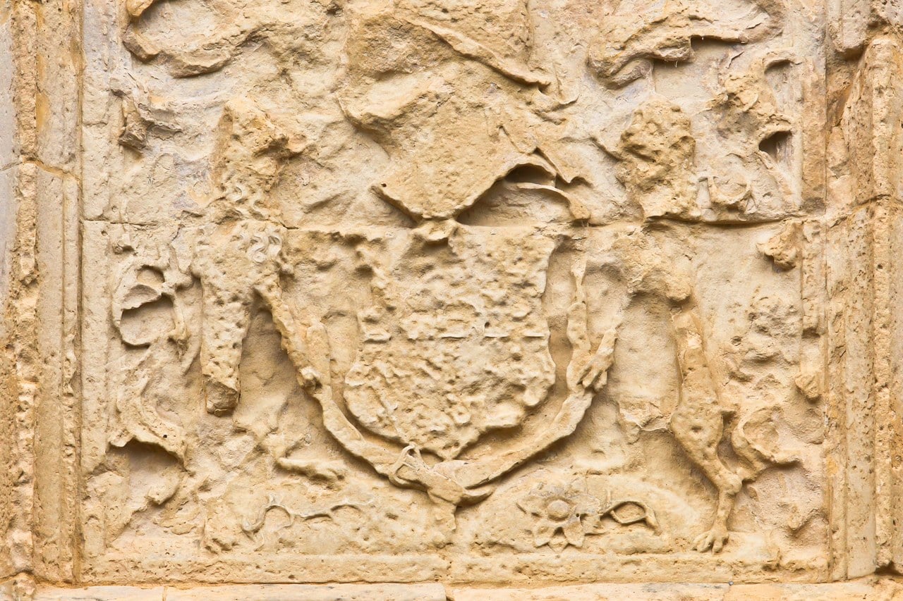 A carved stone panel set in a wall, weathered and worn. Within a molded rectangular border, a shield sits in the center carrying a quartered design too eroded to read clearly, flanked by two standing animal supporters carved in relief. A carved ornament runs along the base beneath the shield. The stone is pale and pitted. A carved stone panel set in a wall, weathered and worn. Within a molded rectangular border, a shield sits in the center carrying a quartered design too eroded to read clearly, flanked by two standing animal supporters carved in relief. A carved ornament runs along the base beneath the shield. The stone is pale and pitted.