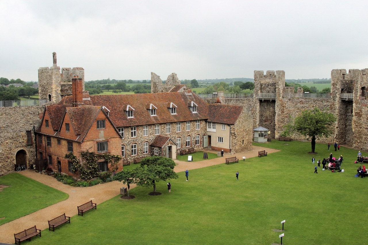 The inner court of Framlingham Castle seen from the wall-walk. A broad level lawn fills the center of the frame, with benches and visitors on it and a gravel path curving round. Along the left stands a long two-storey building with a red tiled roof, dormer windows and tall chimney stacks, its left end built of red brick and the rest of pale flint and stone. The gray curtain wall with square towers runs round the far side of the court, with open countryside beyond. The inner court of Framlingham Castle seen from the wall-walk. A broad level lawn fills the center of the frame, with benches and visitors on it and a gravel path curving round. Along the left stands a long two-storey building with a red tiled roof, dormer windows and tall chimney stacks, its left end built of red brick and the rest of pale flint and stone. The gray curtain wall with square towers runs round the far side of the court, with open countryside beyond.