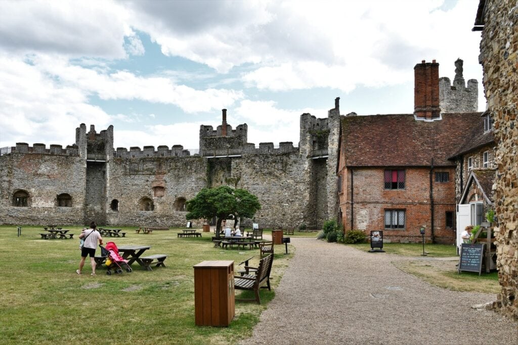 Framlingham Castle 7 The inner court of the castle on a bright day. Level mown grass fills the foreground with wooden picnic benches on it and visitors walking, including an adult pushing a pushchair. The crenellated flint curtain wall runs across the background with square towers along it and arched recesses in its inner face. On the right stands a red brick building with a tall chimney and a modern glazed doorway. Blue sky with white cloud.