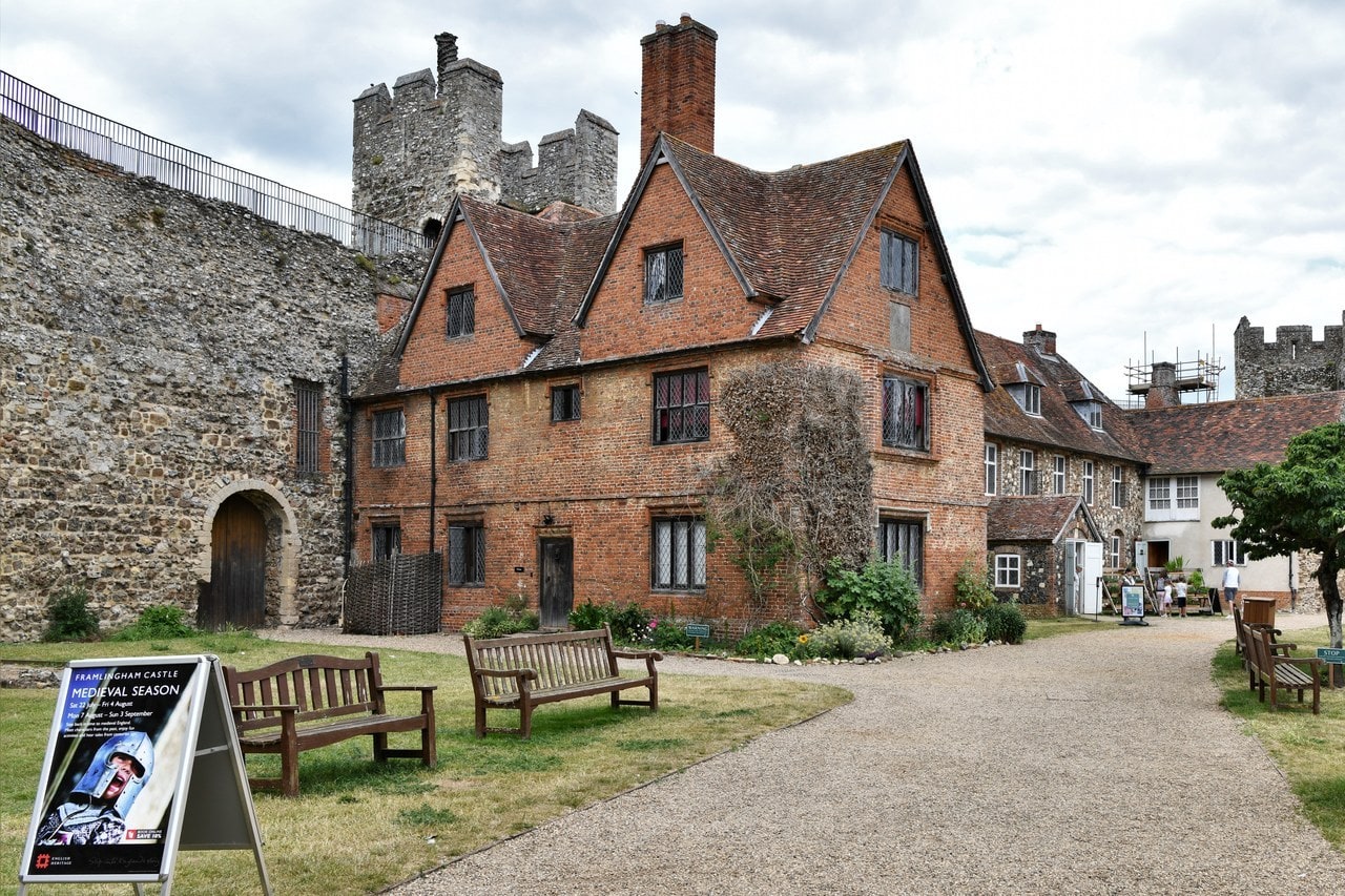 A two-storey red brick building standing inside the castle walls. It has a steep red tiled roof with gabled dormers and two tall chimney stacks, and lower ranges adjoin it to the right. The gray flint curtain wall runs behind it on the left, with an arched opening at its foot, and stone towers rise behind the roofline. Wooden benches and a gravel path stand on the grass in front, with a small display board at the lower left. A two-storey red brick building standing inside the castle walls. It has a steep red tiled roof with gabled dormers and two tall chimney stacks, and lower ranges adjoin it to the right. The gray flint curtain wall runs behind it on the left, with an arched opening at its foot, and stone towers rise behind the roofline. Wooden benches and a gravel path stand on the grass in front, with a small display board at the lower left.