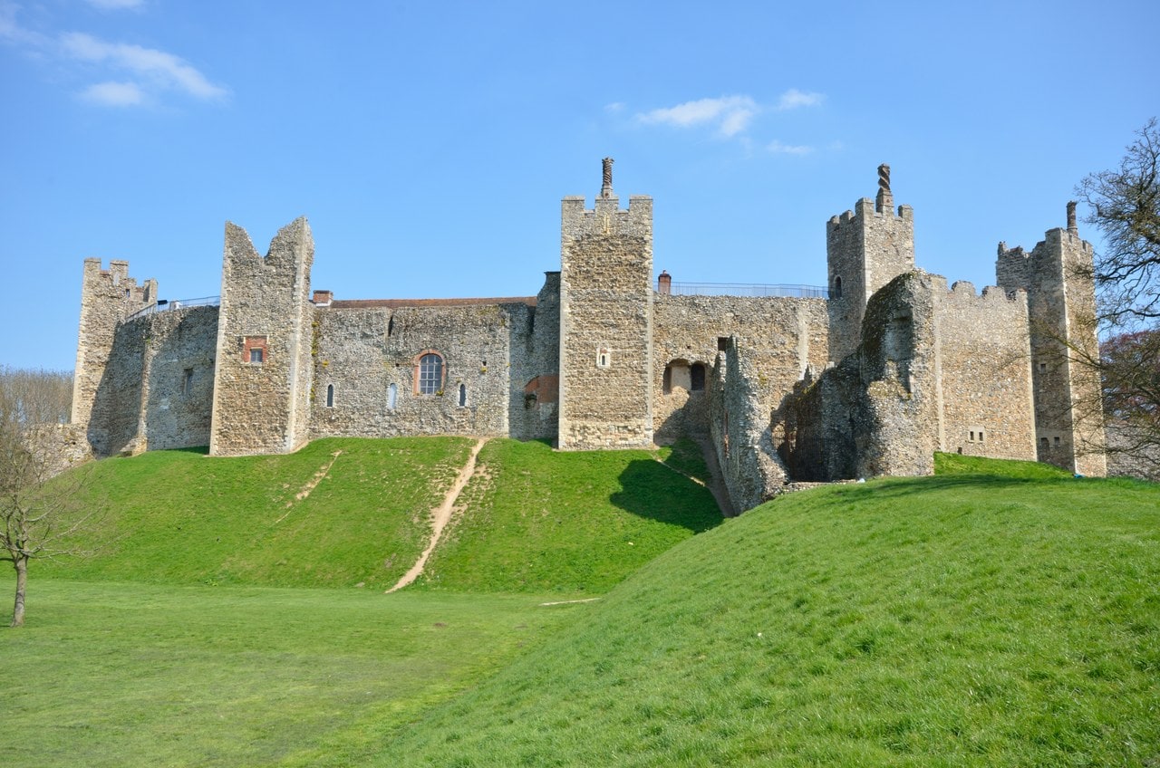 The south side of Framlingham Castle seen from below on a sunny day. A long flint and stone curtain wall runs across the frame with tall square towers rising at intervals along it, two of them carrying slender chimney shafts. A broad bank of mown grass rises steeply from the foreground to the foot of the wall, with a worn footpath climbing it. The sky is clear blue. The south side of Framlingham Castle seen from below on a sunny day. A long flint and stone curtain wall runs across the frame with tall square towers rising at intervals along it, two of them carrying slender chimney shafts. A broad bank of mown grass rises steeply from the foreground to the foot of the wall, with a worn footpath climbing it. The sky is clear blue.