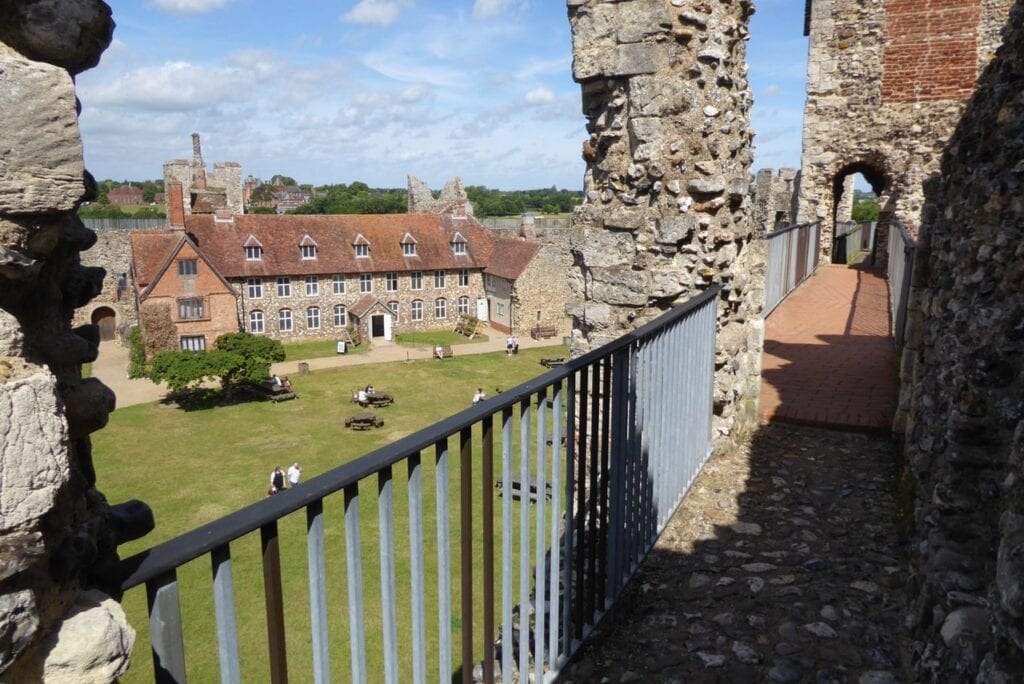 Framlingham Castle 5 The wall-walk along the top of the castle curtain, paved in red brick with a black metal railing running along its left side. Rough flint masonry stands at the right and in the foreground. Ahead the walkway continues toward an arched opening through a tower. Below and to the left lie the green inner court and the long tiled-roofed range, with visitors on the grass. Blue sky with light cloud.