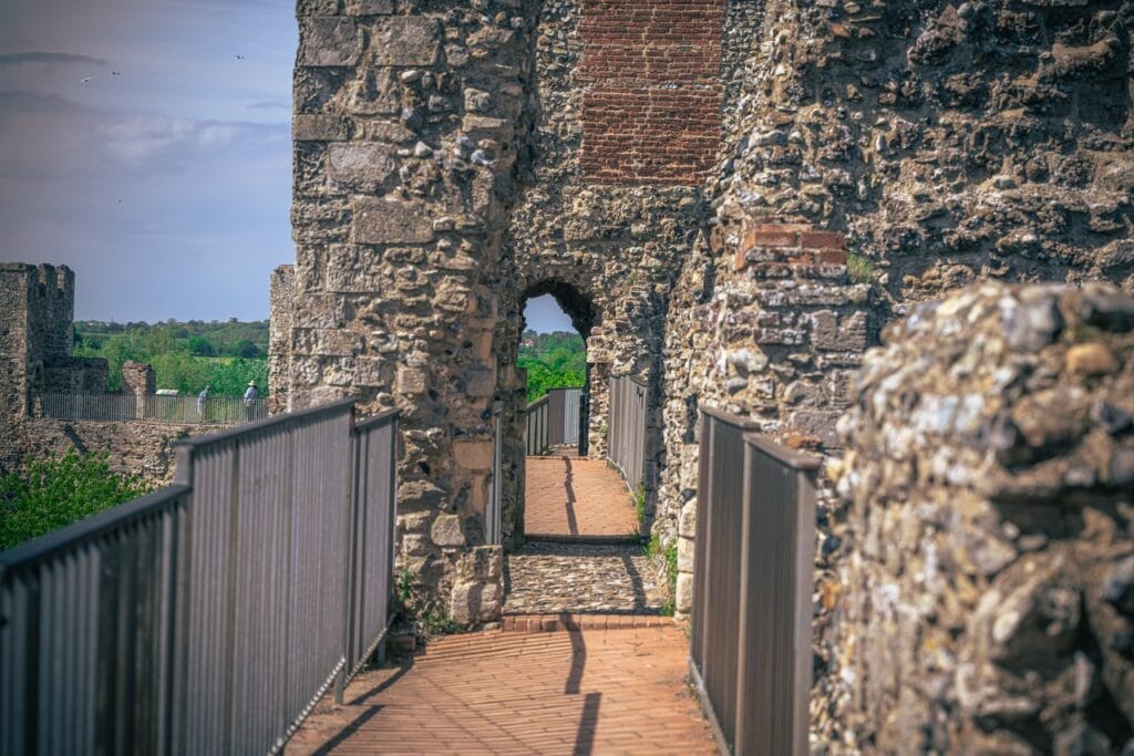 Framlingham Castle 6 A walkway paved in red brick running along the top of the castle wall, with metal railings on the left and rubble masonry rising on the right. Ahead the walkway passes through a narrow arched opening in a tower, with red brick visible in the masonry above the arch. Beyond and to the left, a crenellated stretch of wall, green trees and a strip of water can be seen in the distance.